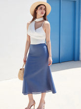 Woman wearing a white top and blue skirt with a straw hat against a blue wall.